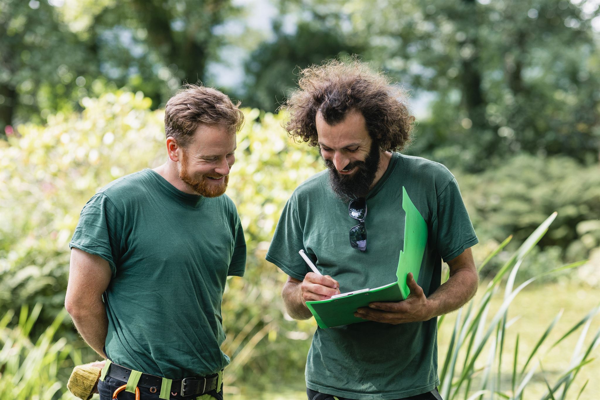 Two-men-in-work-T-shirts-standing-outdoors,-with-grass,-shrubs,-and-trees-behind,-looking-at-folder-contents-held-by-one-man